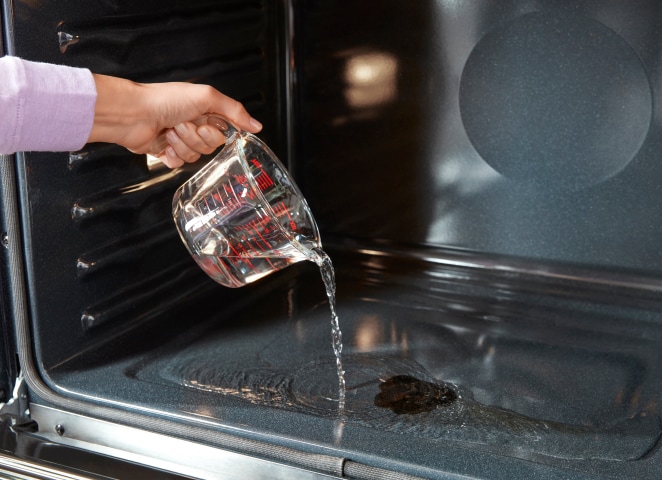 A hand pouring water from a glass measuring cup onto the bottom of a wall oven