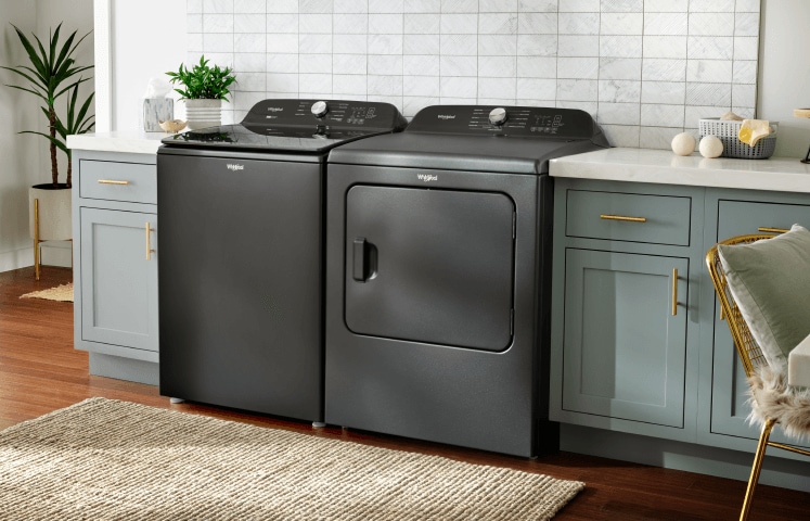 A Volcano Black top load Whirlpool® Washer and Dryer in a laundry room with light cabinets