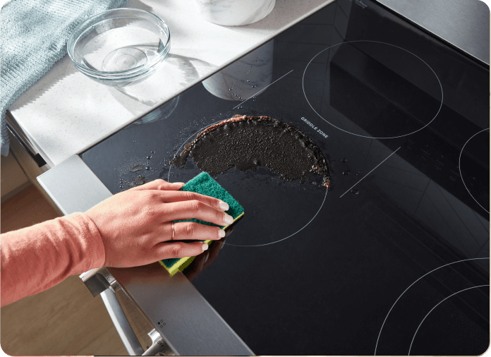 Person wiping baked-on foods from the cooktop with a sponge.