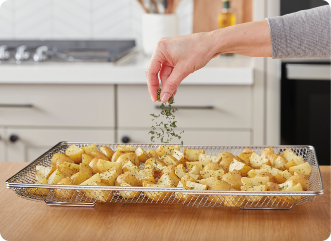 A hand sprinkling herbs onto air fried potatoes in an air fry basket